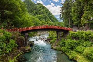 Wide angle view of a japanese landscape with green sloping hills, a red wooden bridge over a brook, against a blue sky with clouds