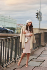 Young pretty beautiful stylish woman in pink dress, white beret hat and beige coat with white leather handbag standing and posing at city street. Fashionable stylish autumn look in pastel colors