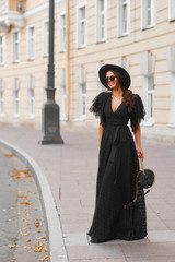 Fototapeta premium Elegant woman in long black dress, hat and sunglasses holding handbag standing and posing at city street. Stylish fashionable warm autumn look. Full length outdoor portrait