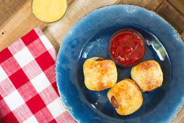 Homemade Mini Pretzel Dogs on a Blue Plate with a Red and White Checked Napkin on a Wooden Tray; Small Bowls of Ketchup and Mustard in Background