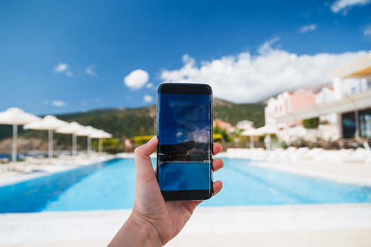 Young woman makes photo of swimming pool area, blue sky and beautiful landscape at background on smartphone camera, to share on internet social media through photo application for mobile device