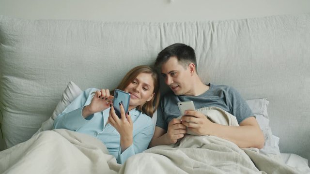 Pretty blonde is using smartphone in bed sharing content with husband smiling enjoying social media together. Modern lifestyle, device and relationship concept.