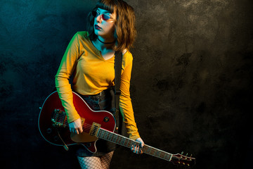Charming hipster woman with curly hair with red guitar in neon lights. Rock musician is playing...