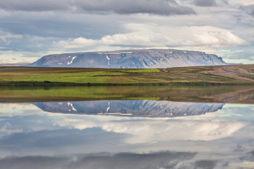 Icelandic landscape with a meadow and a distant mountain reflecting on water under a cloudy sky