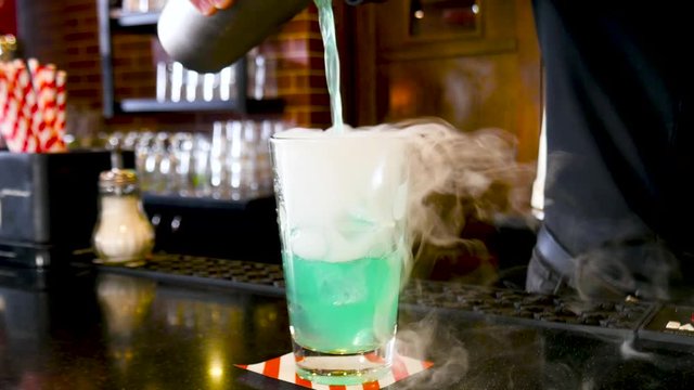 Classic bartender pouring colorful liquid from shaker to a cocktail glass with cold smoke in interior classy bar