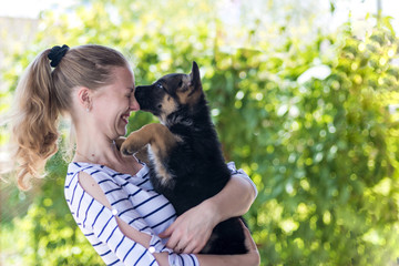 Happy girl holding a German shepherd puppy and smiling. Buying and acquiring a dog, the joy of meeting. The dog bites and kisses the owner. On a blurred green background. copy space