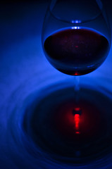 Fototapeta na wymiar Wine glass with red wine and galaxy inside highlighted with flashlight from above. Contrast outline on dark blue rippled background with red light spot.