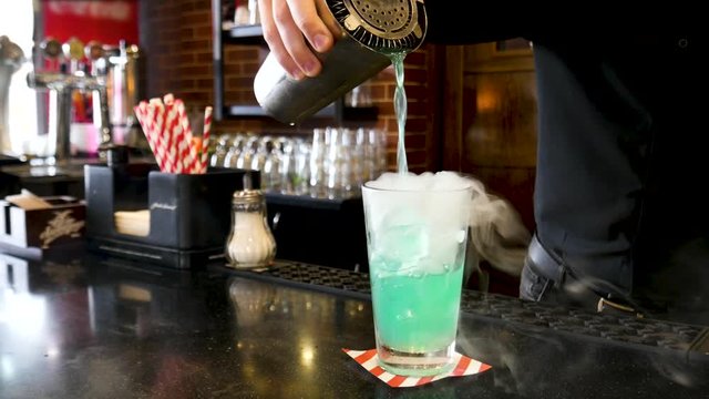 Classic bartender pouring colorful liquid from shaker to a cocktail glass with white cold smoke in interior classy bar