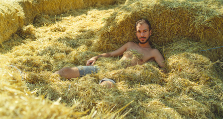A young handsome man with beard of slim build in a t-shirt and shorts is lying in the hay on a...