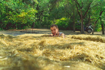A young handsome man with beard of slim build sits in the hay and holds hay in his hands against the background of green trees in the park.