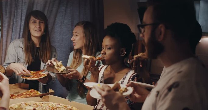 Beautiful young multiethnic women chat, smile eating pizza hanging out with friends at kitchen house party slow motion.