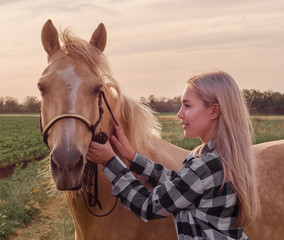 Obraz premium young blonde girl with a beige horse on the ranch on the background of a green field and evening sky