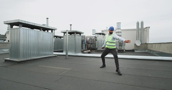 Young constructor man with a beard in a hardhat safety helmet dancing very excited on the rooftop of construction site he feels very happy