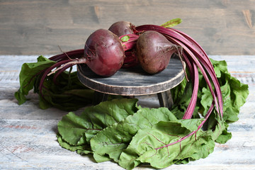 An image of three red beets with leaves lying on a dark round stand, standing on a light wooden surface on a gray wooden background