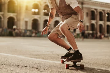 Poster Urban man lifestyle. Stylish man ride on skateboard in white shirt on city street. Portrait of handsome bearded hipster male near road on buildings background. © Yevhen
