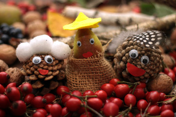 Fototapeta premium Three faces made of cones with eyes and mouth, head cover surounded by falll items as rose hip, nuts, grapes, leaves. Autumn background.