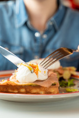 Close up of cutting a poached egg with knife and fork as a part of breakfast with soft waffle, mashed avocado and salmon
