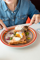 Young woman is cutting a poached egg with knife and fork as a part of breakfast with soft waffle, mashed avocado and salmon