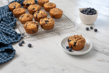 Homemade Blueberry Muffins on Cooling Rack with One Isolated in Front on Small White Plate; Bowl of Blueberries in Background; White Countertop