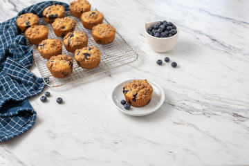 Homemade Blueberry Muffins on Cooling Rack with One Isolated in Front on Small White Plate; Bowl of Blueberries in Background; White Countertop