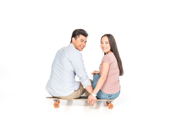 cheerful asian couple sitting on longboard and smiling at camera on white background