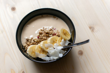 Bowl of granola with yogurt, banana slices and coconut chips on light brown wooden board for healthy breakfast