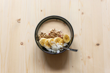 Bowl of granola with yogurt, banana slices and coconut chips on light brown wooden board for healthy breakfast