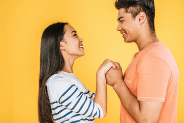 Fototapeta premium happy asian man and woman holding hands while looking at each other isolated on yellow