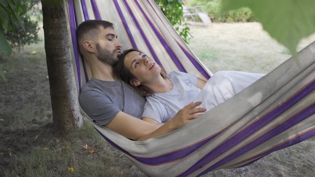 Young caucasian man and woman lying in hammock in the garden relaxing together. Loving couple together outdoors. Summertime leisure