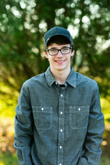 Teenage boy with glasses standing outside in front of a tree wearing a gray button up long sleeved shirt and denim jeans