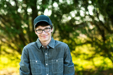 Teenage boy with glasses standing outside in front of a tree wearing a gray button up long sleeved shirt and denim jeans with baseball cap on