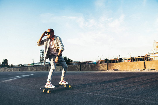 Young caucasian man riding on longboard or skateboard, modern shot in film grain effect and vintage style. Sunset in summer evening. City's street. Toned in teal orange. Looks confident and cool.