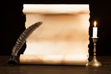 A lit candle and feather with inkwell against the background of an old scroll of parchment