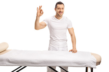 Professional young masseur standing behind a massage bed and gesturing sign good - obrazy, fototapety, plakaty