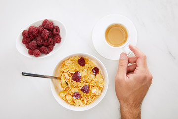 cropped image of man holding cup of coffee near bowl with cornflakes and plate with fresh raspberry on white marble background