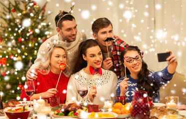 Obraz na płótnie Canvas holidays and celebration concept - happy friends with party props taking selfie by smartphone at home christmas dinner over snow