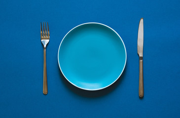 Ready to order. High angle shot of empty color plate, fork, knife lying against blue background.