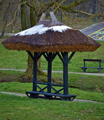 Wooden gazebo covering bench in park, covered by melting snow in spring