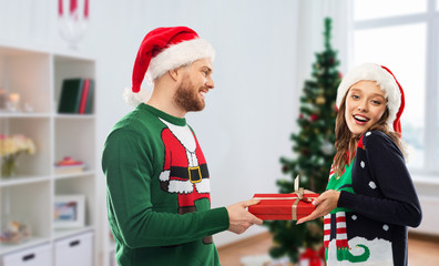 people and winter holidays concept - happy couple in santa hats with gift in ugly sweaters over christmas tree at home background