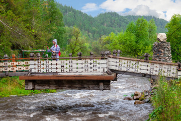 A young blonde girl in a blue raincoat is looking for a path on the navigator on the phone and on the map while traveling through wild places on a wooden suspension bridge over mountain river.