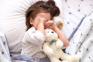 Top view of little girl lying in bed with teddy bear, being in bad mood, does not want to srtand up and go to kinder garten, toddler on pillow rubbing her eyes, looks sad. Childhood concept.