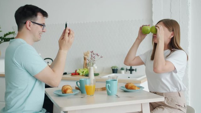 Girl is posing with fruit apples having fun while guy is taking pictures with smartphone smiling sitting at kitchen table together. Family and lifestyle concept.