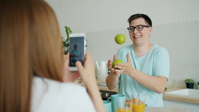 Slow motion of young man juggling apples having fun in kitchen while woman is taking photo with smartphone camera. Gadgets and family lifestyle concept.