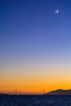 New moon over the Golden Gate Bridge, with an orange sky on the horizon and blue water in the foreground.