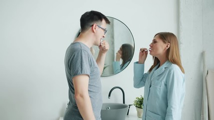 Attractive man and woman in pajamas happy couple are brushing teeth talking smiling in modern apartment. Casual hygiene, relationship and people concept.