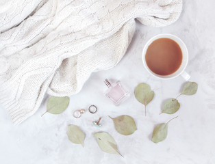 Fashion concept with warm chunky sweater and jewelry next to hot coffee, perfume and eucalyptus leaves. Flat lay. Top view. 