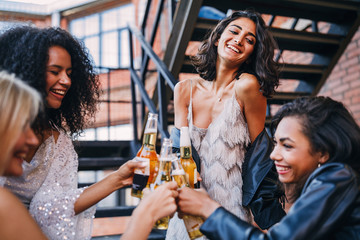 Four cheerful women standing together on the staircase toasting bottles of beer