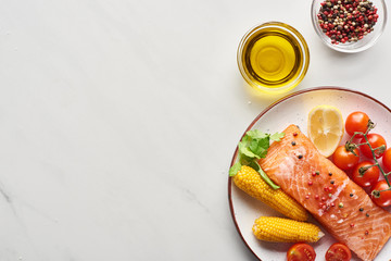 top view of raw salmon steak with corn and tomatoes on plate near oil and peppercorns on marble...