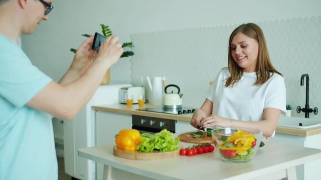 Housewife attractive blonde is making salad having fun while man is taking pictures with smartphone camera. Lifestyle, food and modern technology concept.