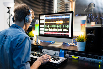 Musician with headphones pressing keys of piano keyboard by computer screen